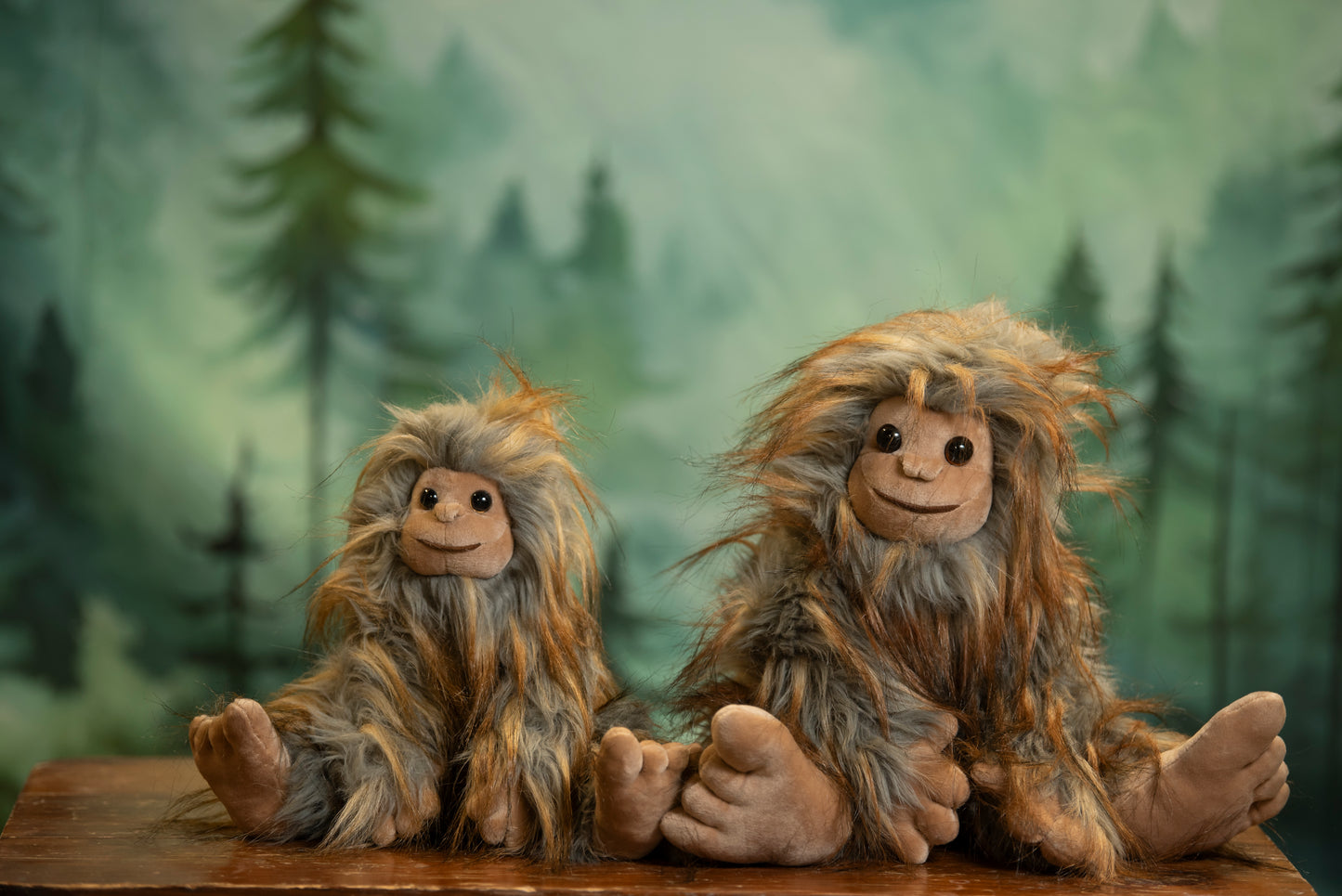 Two plush Sasquatch toys sitting on a table with a forest background