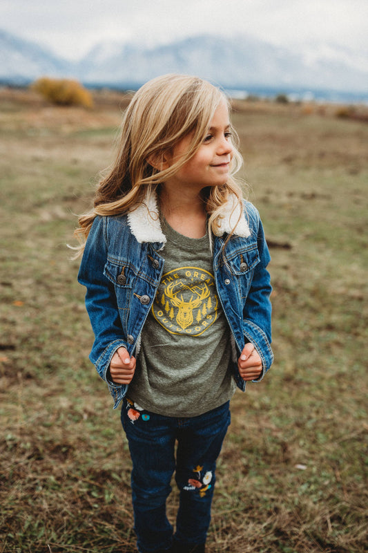 girl wearing olive green t-shirt with yellow graphic with an elk, mountains and starts on it.