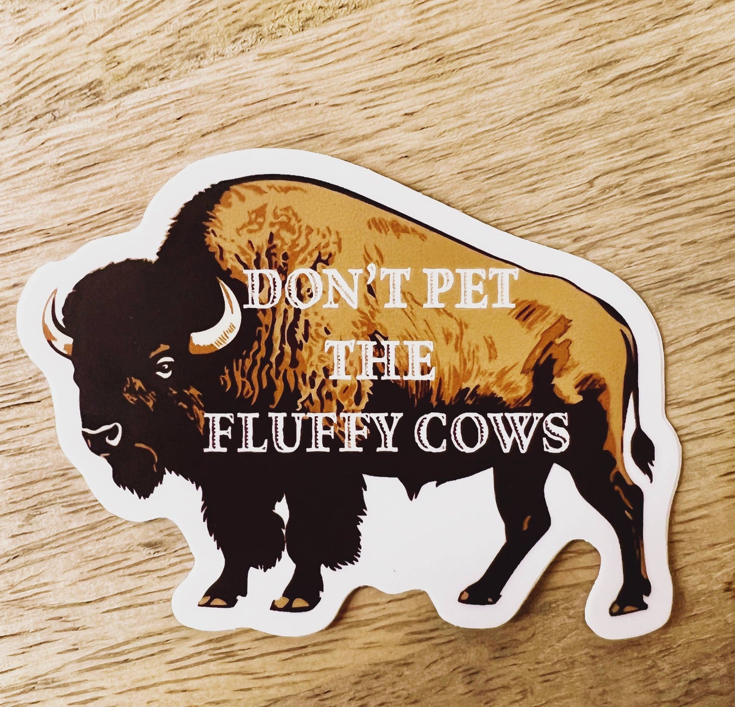 Sticker of a bison with text 'Don't pet the fluffy cows' on a wooden surface