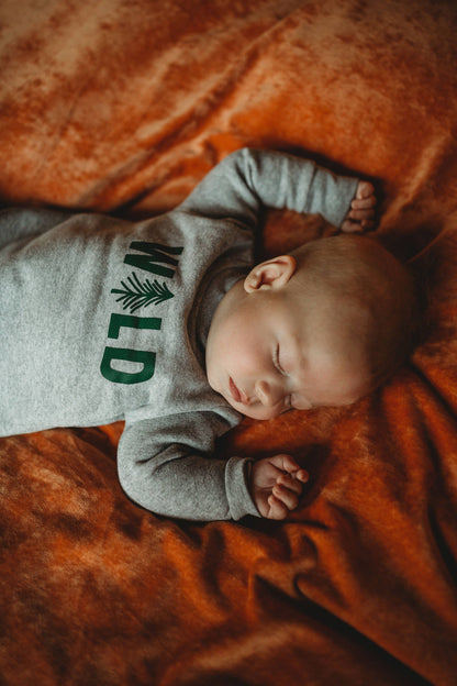 Baby sleeping on an orange blanket wearing a gray sweatshirt with 'WILD' printed on it.