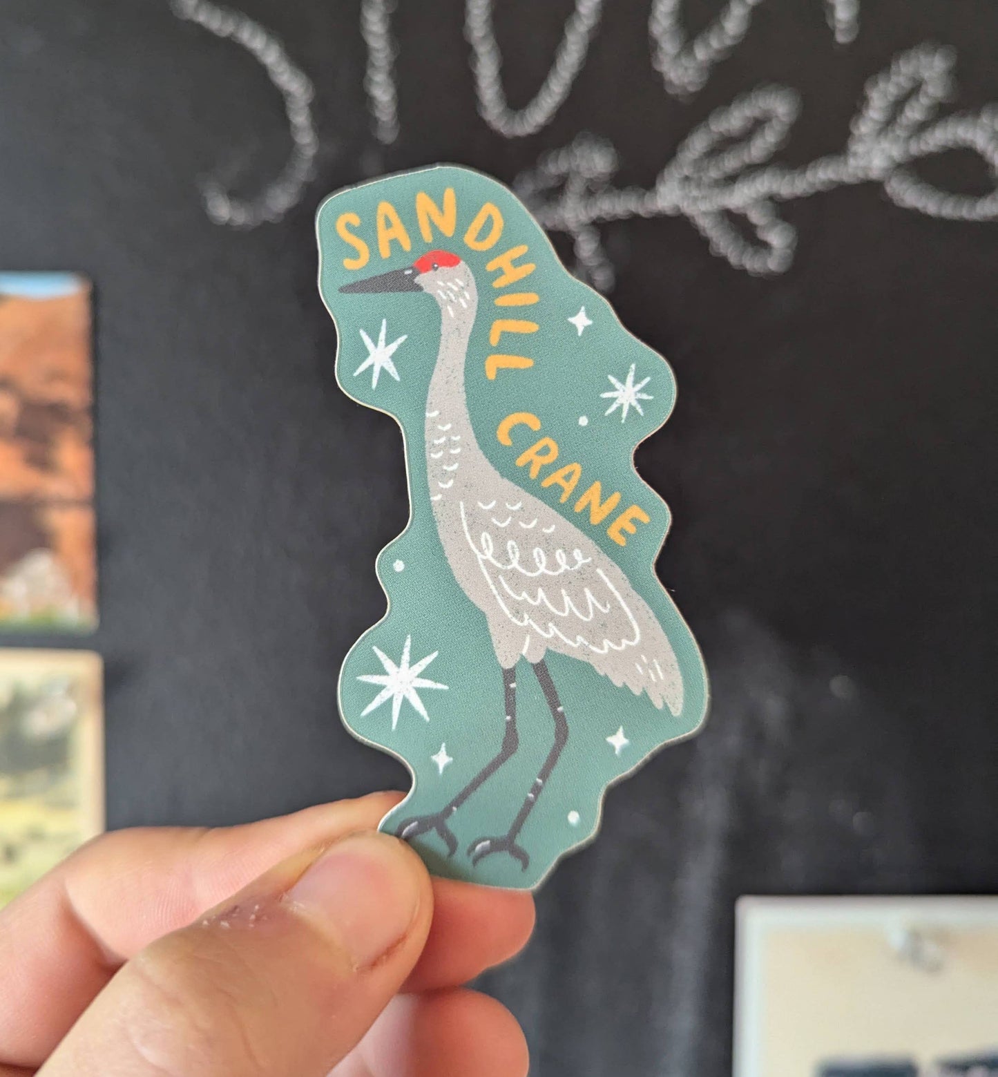 Sticker of a sandhill crane held by a hand against a chalkboard background