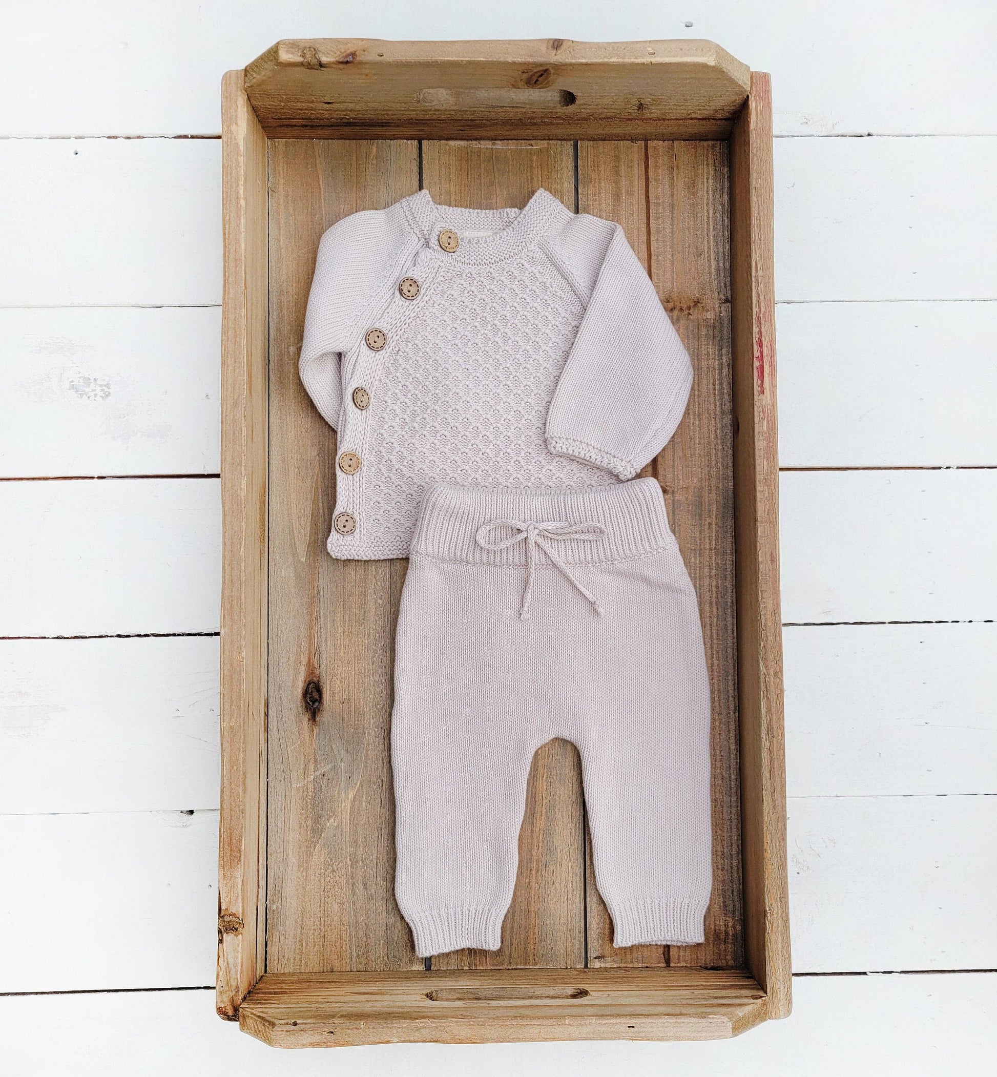 Knitted baby outfit with cardigan and pants in a wooden box on a white wooden floor.