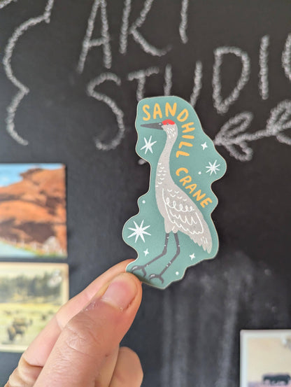 Hand holding a sandhill crane sticker against a chalkboard background with 'Art Studio' written on it.