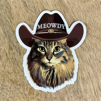 Cat sticker with a cowboy hat labeled 'MEOWDY' on a wooden surface