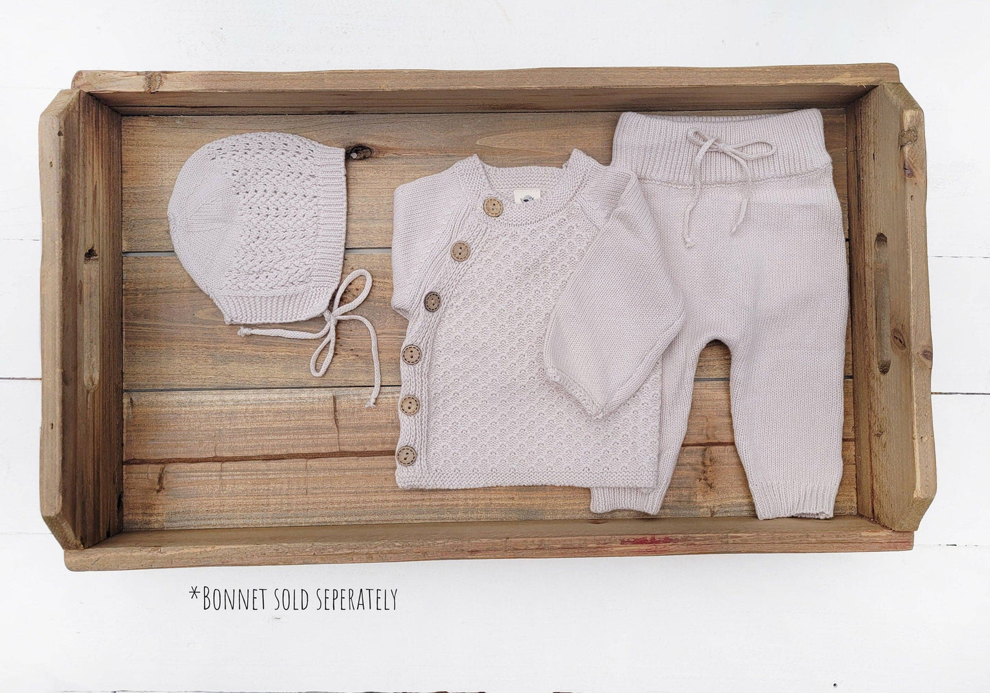 Baby outfit set with cardigan, pants, and bonnet on a wooden tray.