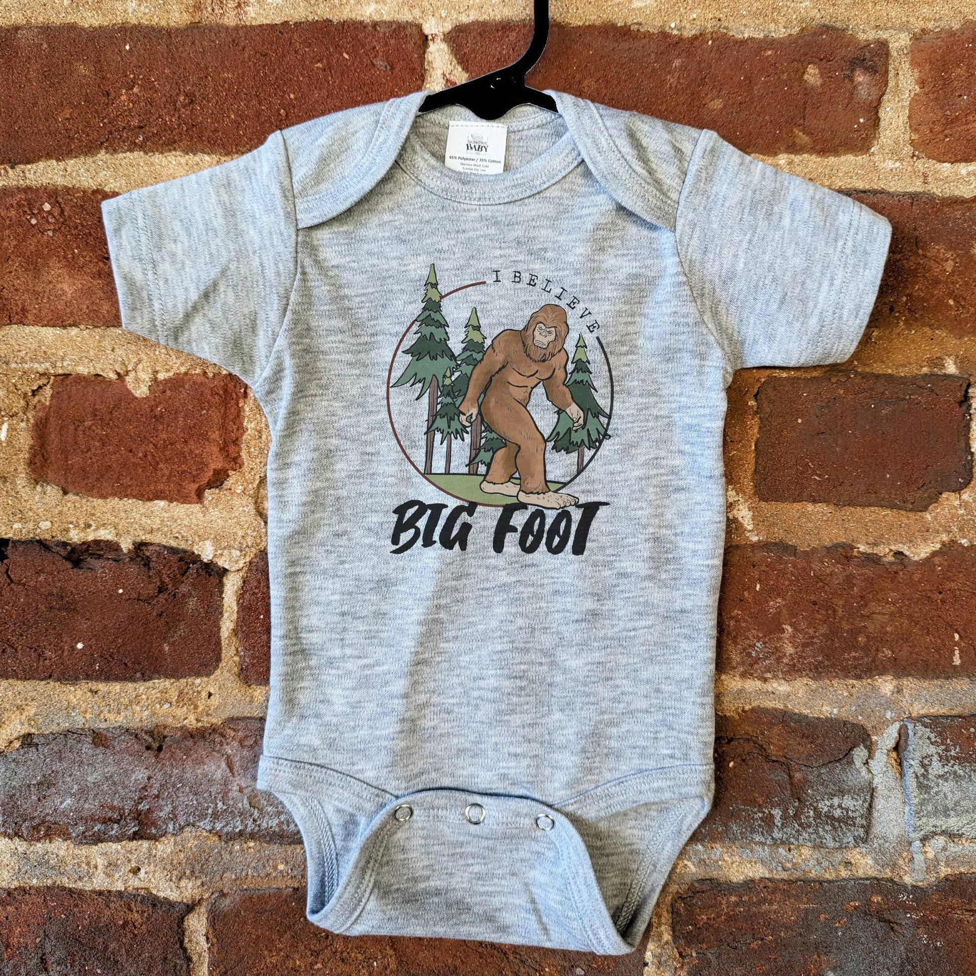 Gray baby onesie with Bigfoot graphic on a brick wall