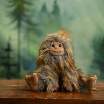 Plush toy resembling a creature with long hair on a wooden surface with a forest background