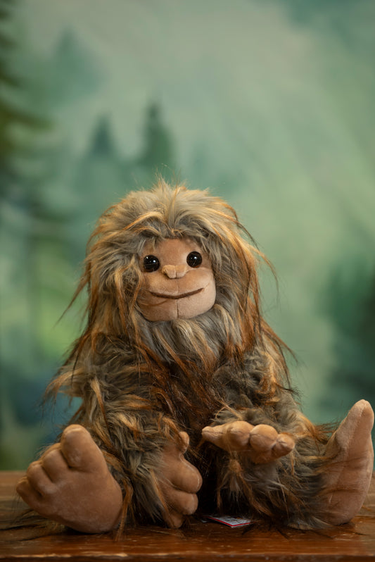 Plush toy resembling a creature with long hair and a beard, sitting on a wooden surface with a blurred forest background.