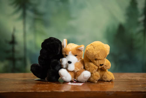 Three stuffed animals (black, white, and brown) sitting together on a wooden surface with a forest background.