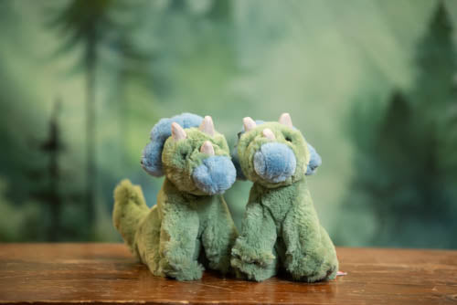 Two green plush dinosaurs with blue faces on a wooden surface with a blurred forest background