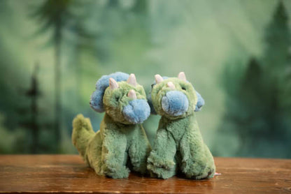 Two green plush dinosaurs with blue faces on a wooden surface with a blurred forest background
