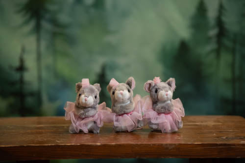 Three stuffed animal toys in pink dresses on a wooden surface with a forest background