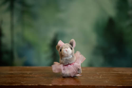 Small plush mouse toy in a pink dress on a wooden surface with a blurred green background