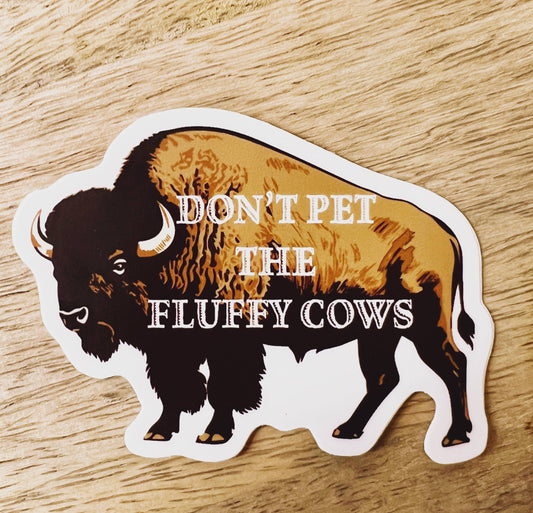 Sticker of a bison with text 'Don't pet the fluffy cows' on a wooden surface