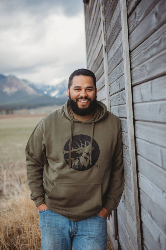 Man wearing a green hoodie with a logo, standing next to a wooden building with mountains in the background.