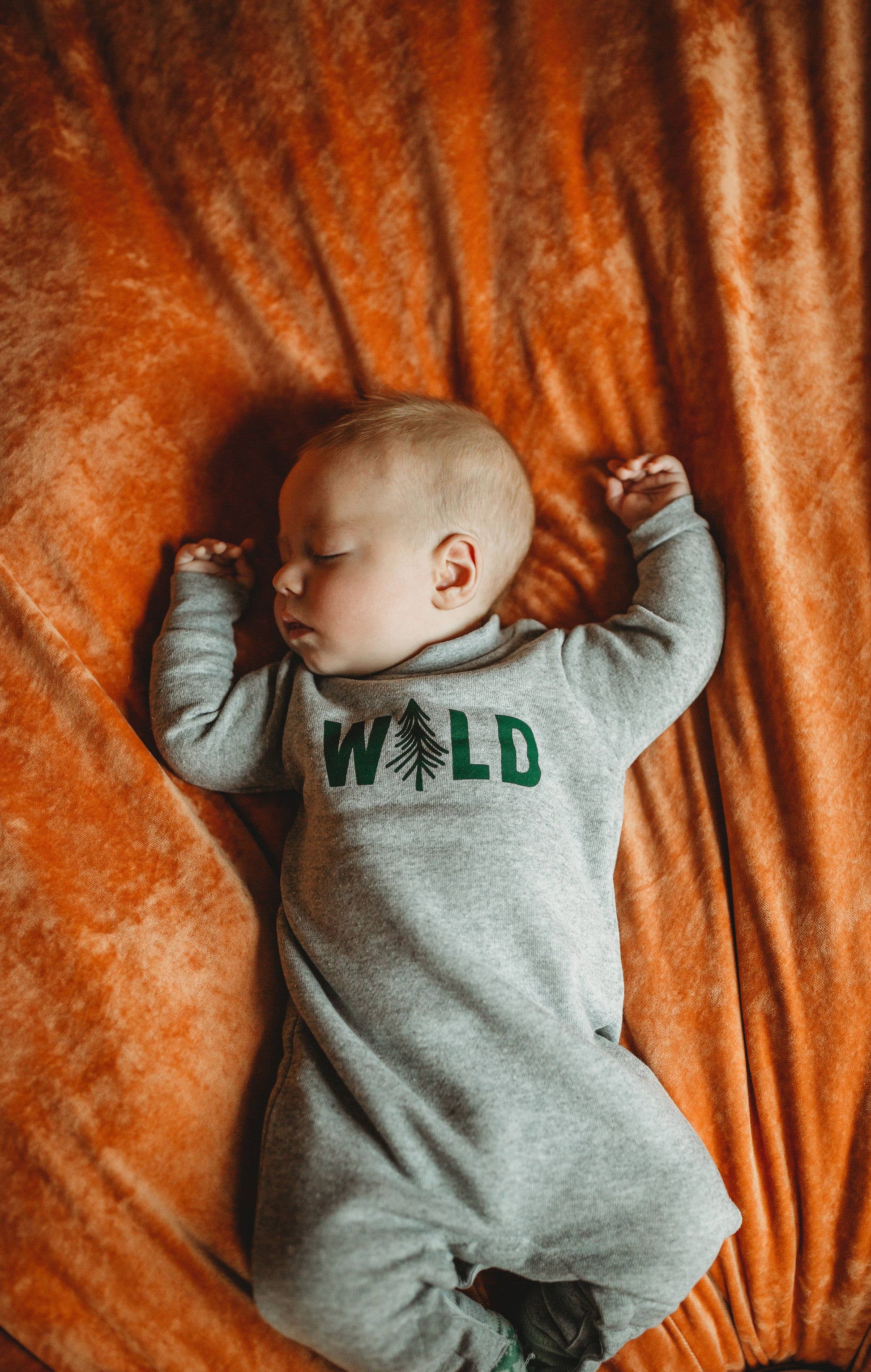 Newborn baby wearing a gray onesie with 'WILD' and tree graphics, lying on an orange blanket.